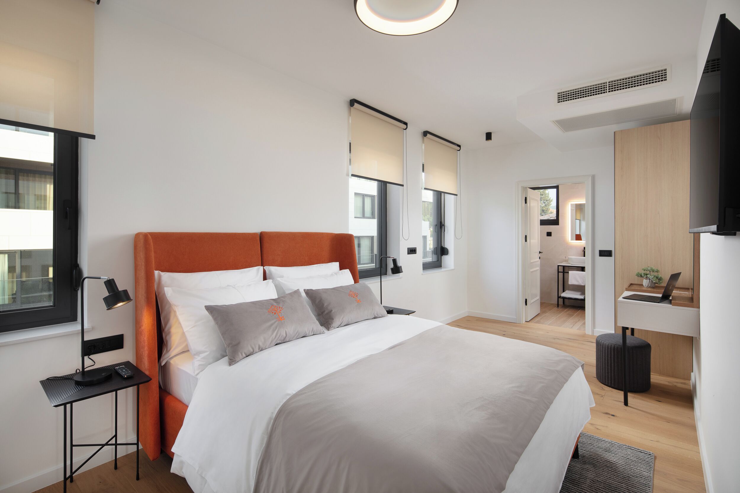 Quarto de hotel moderno com uma cama grande, uma mesa com um laptop, um espelho e uma janela com persianas.