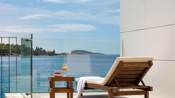 Un balcón con puerta de cristal, una silla de lounge de madera, una mesita con un frutero y un vaso de jugo de naranja, y vista al mar y la costa lejana.