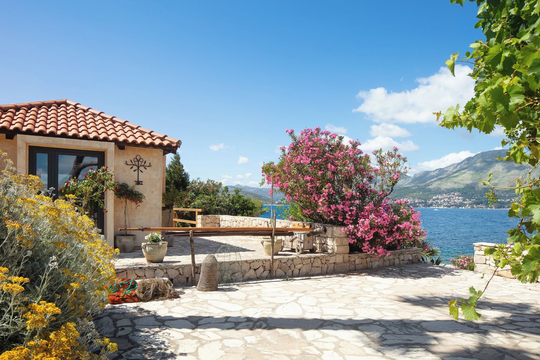A stone patio with a view of a lake and mountains, a building with a tiled roof, and flowering plants.
