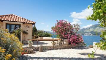 A stone patio with a view of a lake and mountains, a building with a tiled roof, and flowering plants.