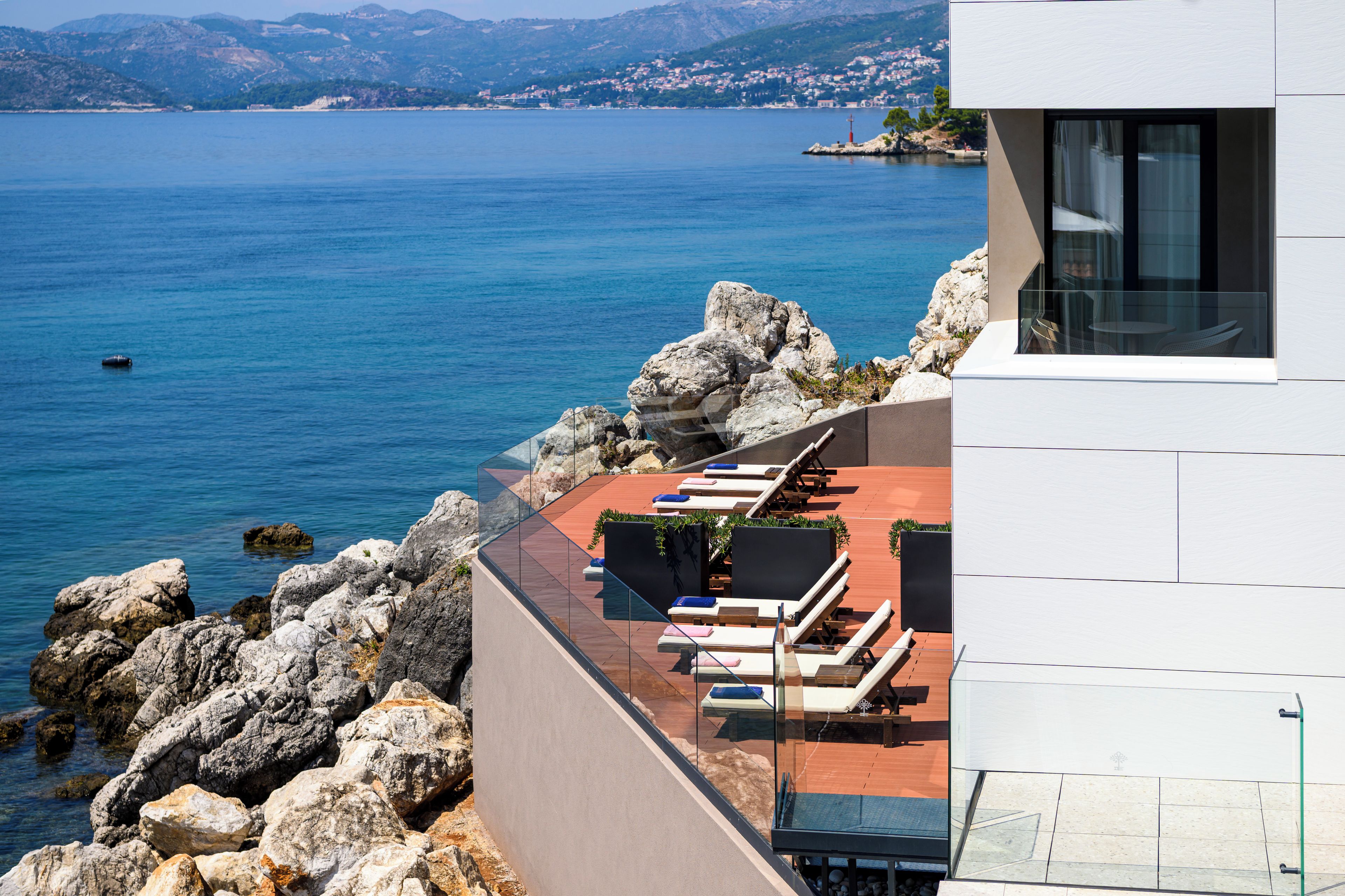 Un bâtiment moderne doté d’une terrasse donnant sur une étendue d’eau, avec des espaces de détente en plein air et une vue sur la côte.
