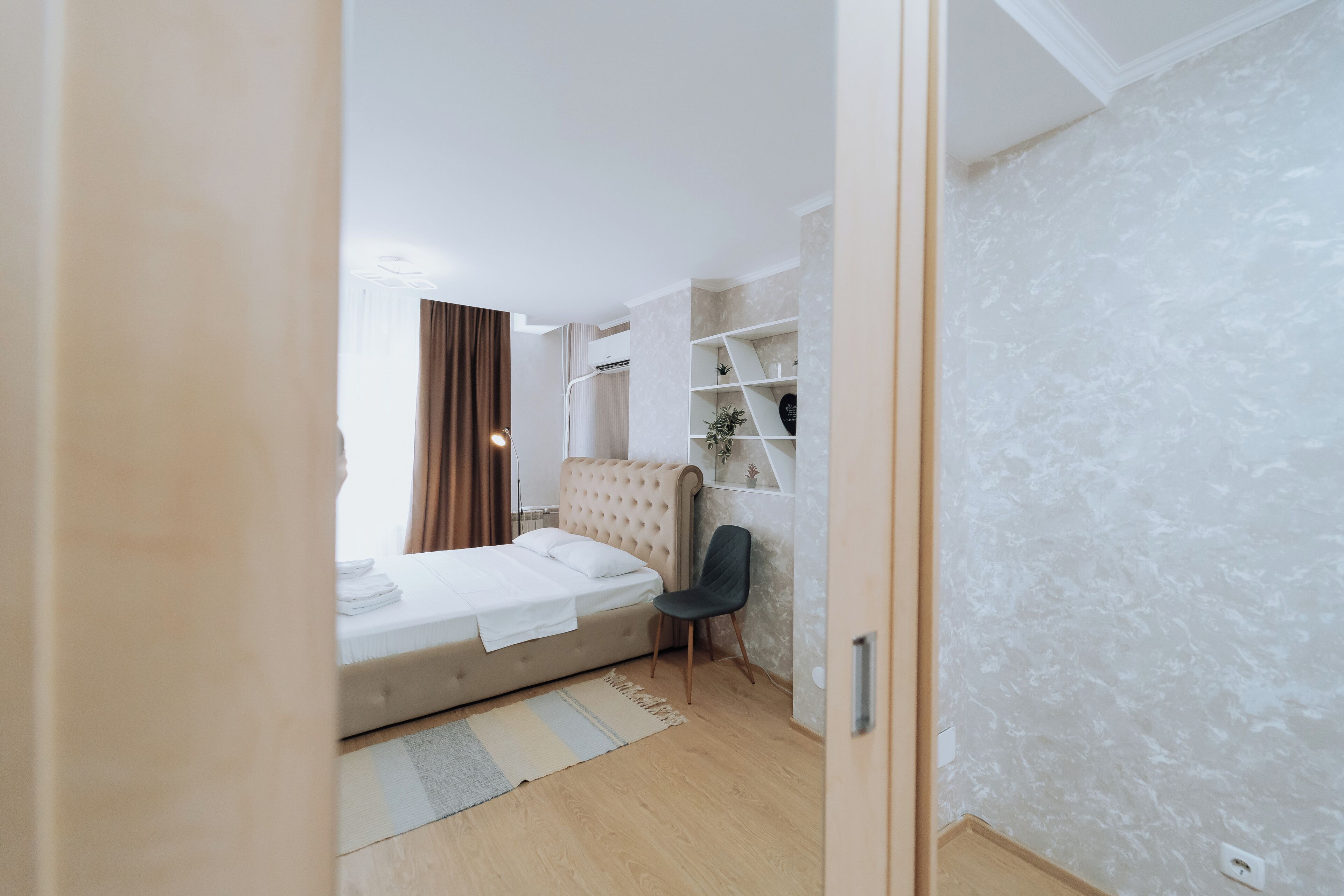 A bedroom with a bed, a chair, a shelf with decorative items, and a window with curtains.