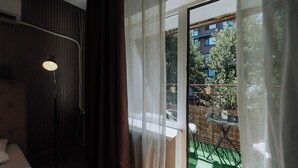 A hotel room with a large window, a bed, a floor lamp, and a small balcony with a table and chairs.