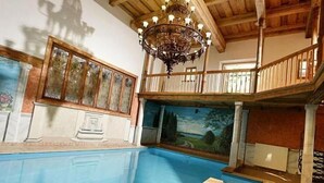 Una piscina interior con techo de madera y candelabro, rodeada por un balcón con vistas a un mural.