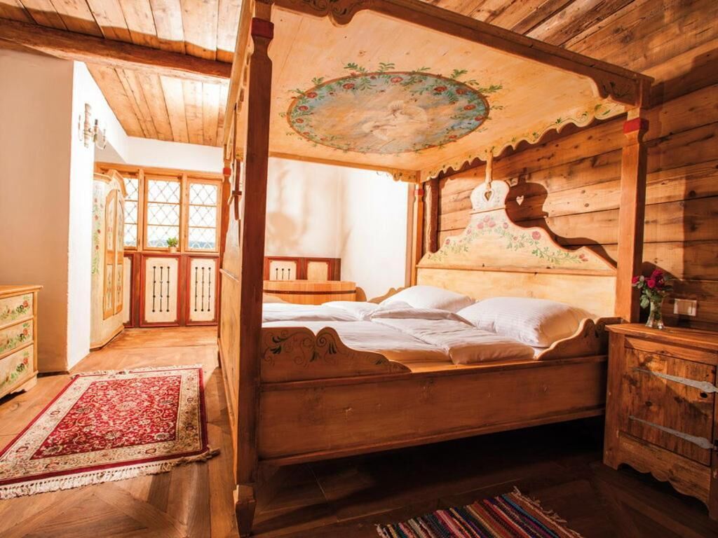 A wooden four-poster bed with a floral ceiling design, a red patterned rug, and a wooden nightstand.