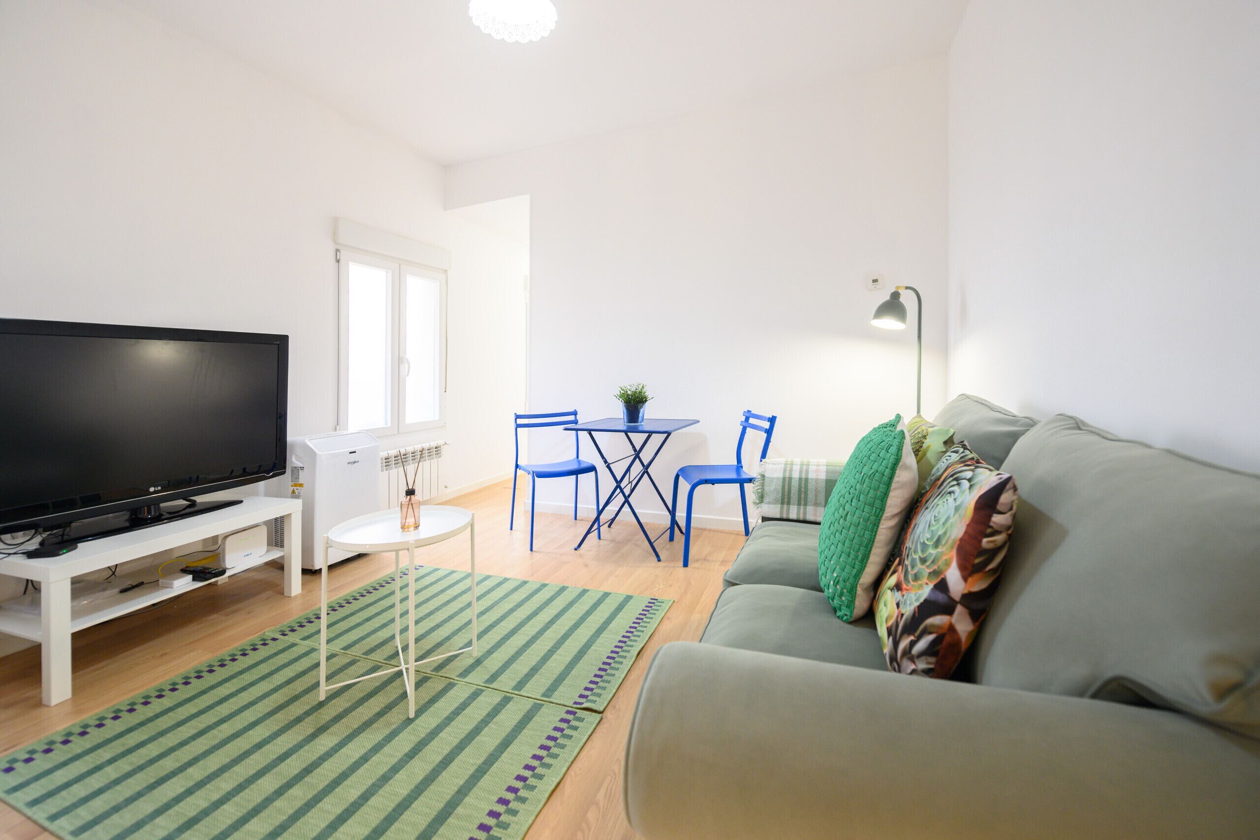 Una sala de estar con una televisión de pantalla plana, un sofá, una mesa de centro y un comedor con sillas y una mesa pequeña.