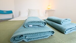 A neatly made bed with folded blue towels and a green bedspread in a simple room with white walls and a nightstand.