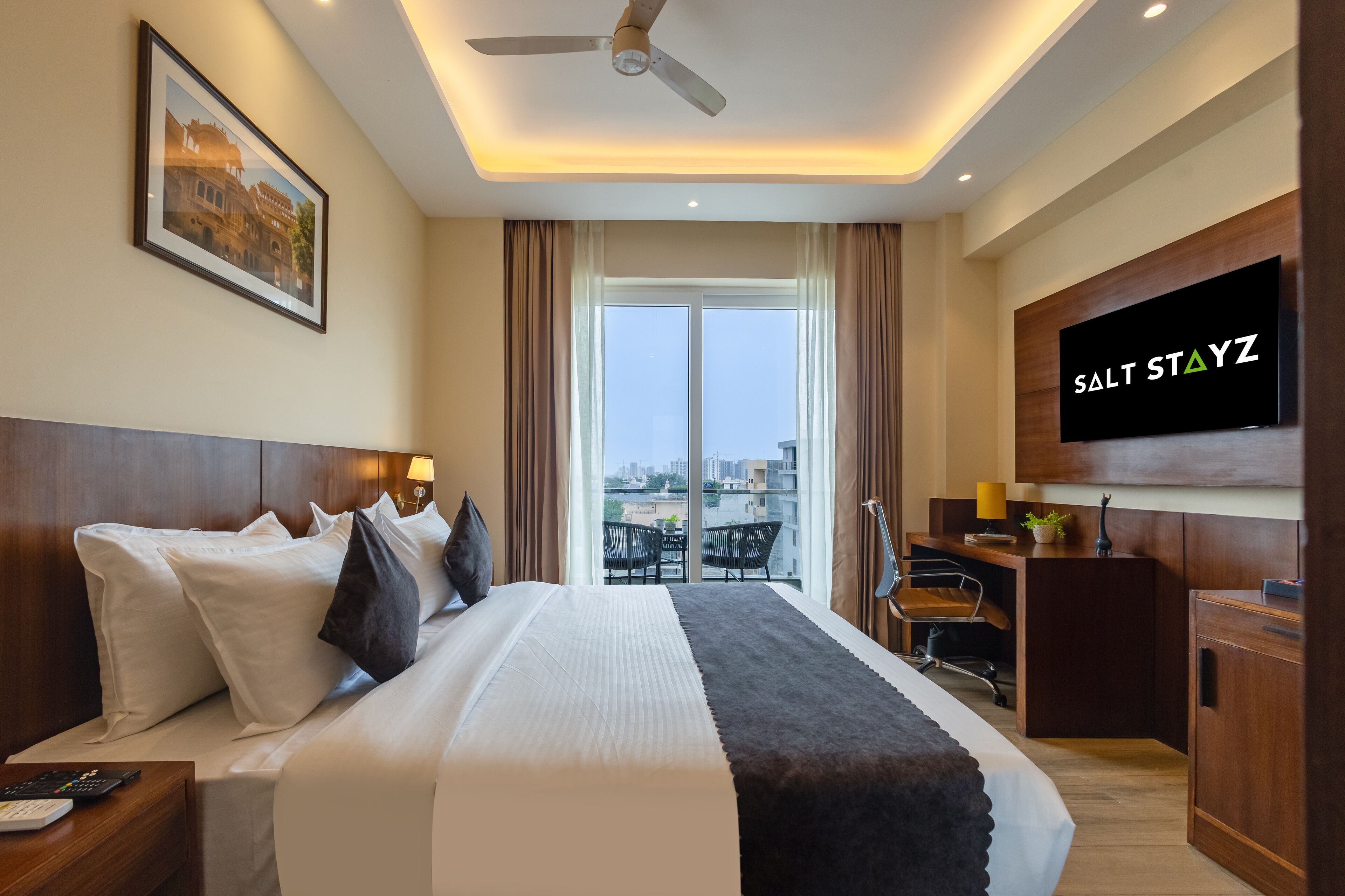 A hotel room with a large bed, a desk with a TV, and a view of the city through the window.