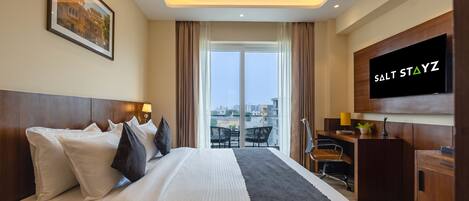 A hotel room with a large bed, a desk with a TV, and a view of the city through the window.