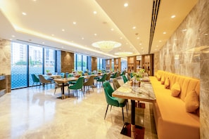 Comedor moderno de hotel con suelos de mármol, sillas verde y beige, un sofá largo color naranja y grandes ventanales con vistas a la ciudad.