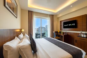 A hotel room with a bed, a TV, a desk, and a view of the city through the window. - Saltstayz Premier - Manesar & Sector 86 (Gurugram)