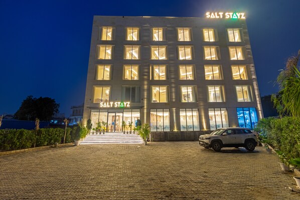 A building with the sign "SALT STAYZ" at the top, featuring multiple illuminated windows and a paved entrance area with a parked vehicle. - Saltstayz Premier - Manesar & Sector 86 (Gurugram)
