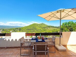 A terrace with a table and chairs, an umbrella, and a view of a green hill and distant buildings. - Bakkehuset by Interhome (Saint Cyr sur Mer La Madrague)