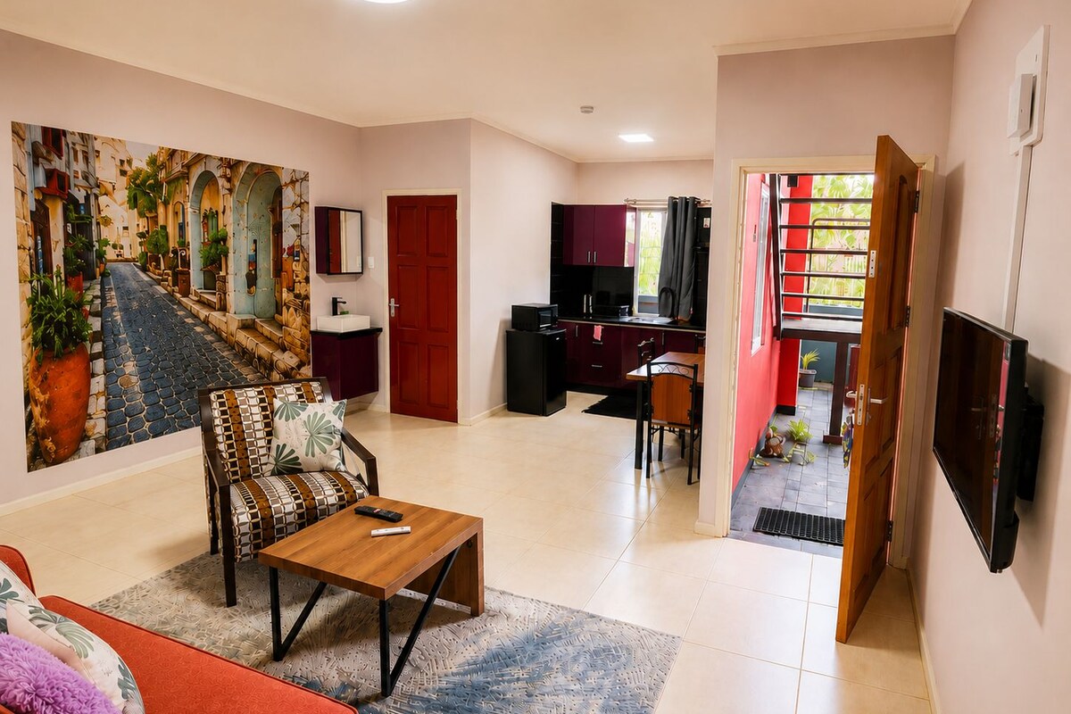 Un salon comprenant un canapé, une table basse, un tableau, un coin repas avec une table et des chaises, un coin cuisine et un téléviseur fixé au mur.