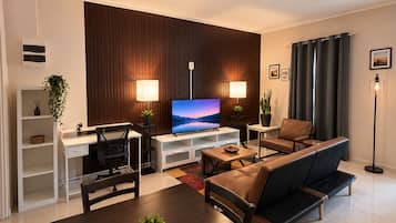 A modern living room with a flat-screen TV, a sofa, a dining table, and a workspace with a desk and chair.