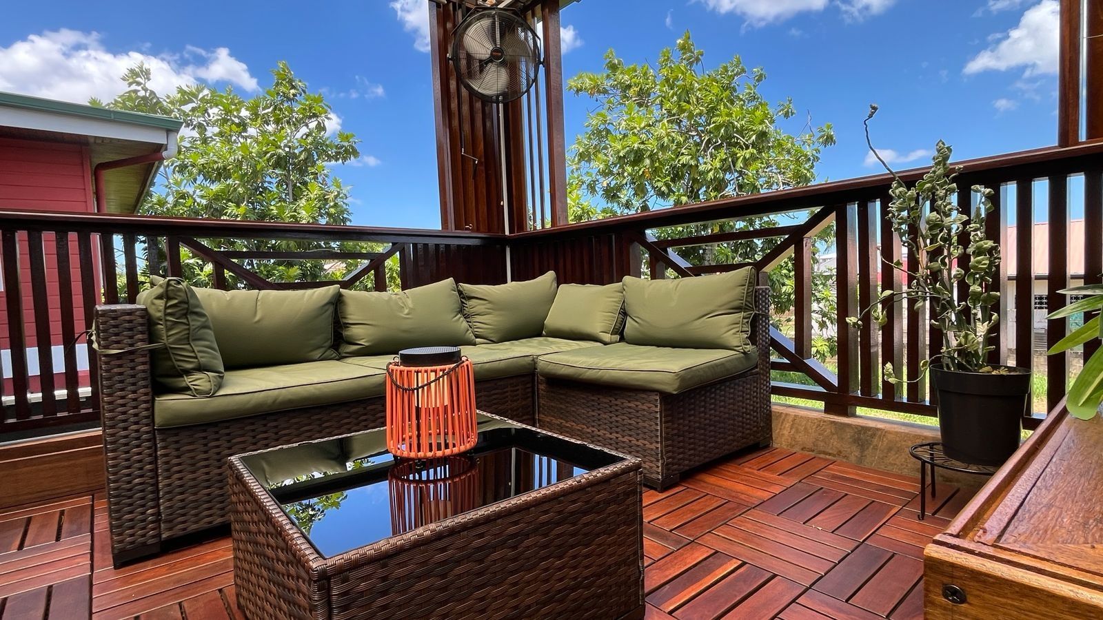 An outdoor deck with a wicker sofa, a glass-top coffee table, and a potted plant.