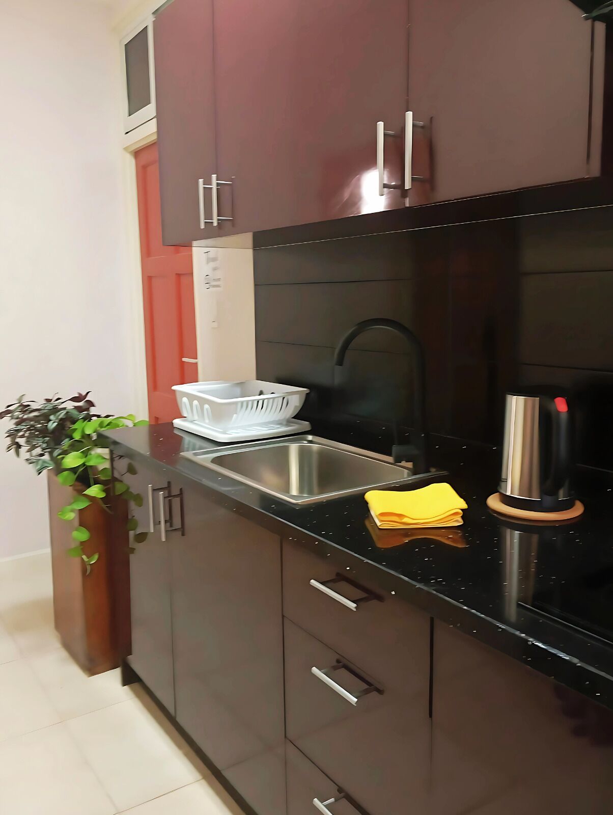 Una cocina moderna con armarios oscuros, un lavamanos de acero inoxidable, una tetera y una planta.