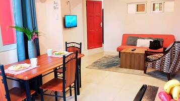 Uma área de refeições com mesa e cadeiras, uma porta vermelha, um sofá, uma televisão e uma pequena planta.