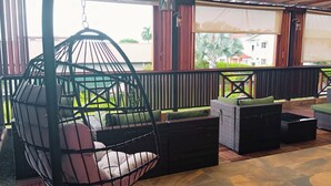 A covered outdoor seating area with wicker furniture, a hanging chair, and a view of a residential area. - Dolfijn Studios (Paramaribo)