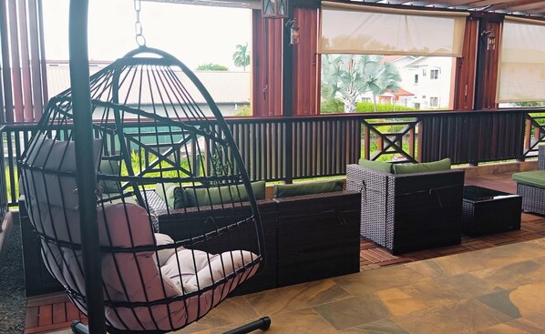 A covered outdoor seating area with wicker furniture, a hanging chair, and a view of a residential area. - Dolfijn Studios (Paramaribo)