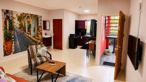 A living room with a sofa, a coffee table, a painting, a dining area with a table and chairs, a kitchen area, and a TV mounted on the wall. - Dolfijn Studios (Paramaribo)