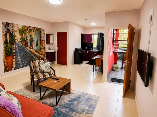 Un salon comprenant un canapé, une table basse, un tableau, un coin repas avec une table et des chaises, un coin cuisine et un téléviseur fixé au mur.