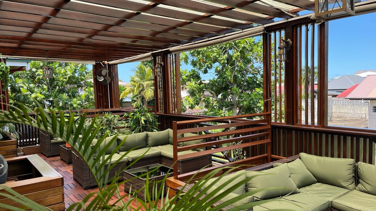 A covered outdoor seating area with a wooden bench and a cushioned sofa, surrounded by greenery.