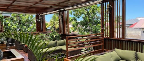 A covered outdoor seating area with a wooden bench and a cushioned sofa, surrounded by greenery.
