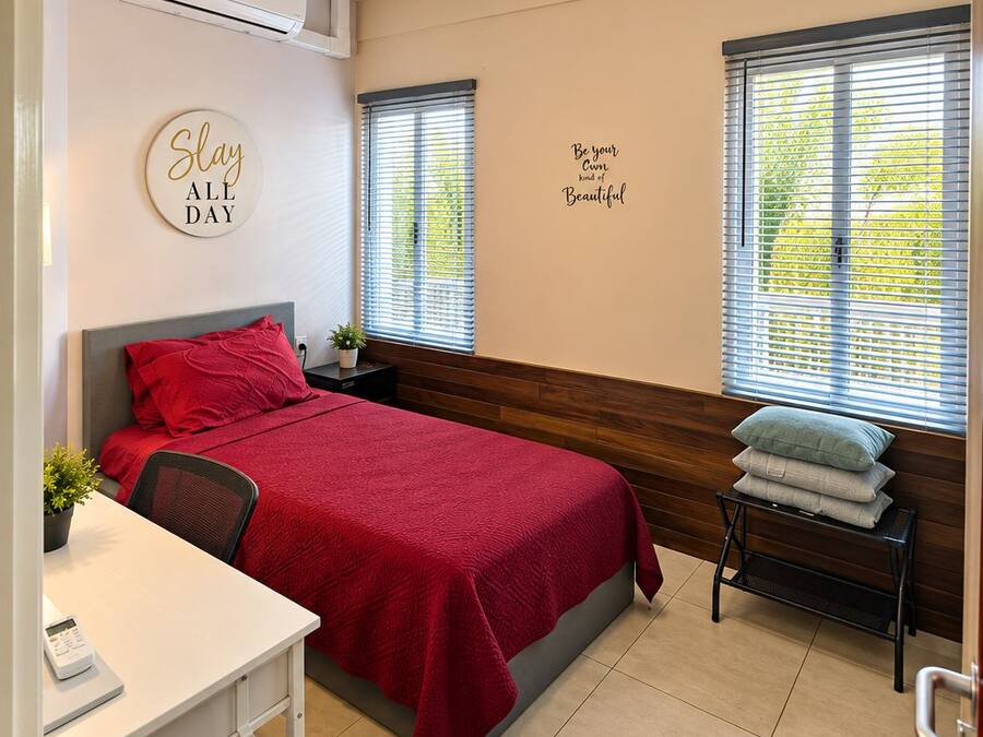 A hotel room with a bed, a desk, a chair, an air conditioning unit, and two windows with blinds.