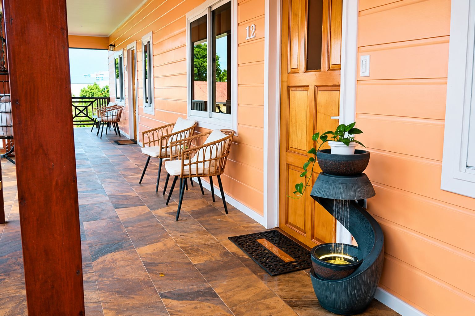 A covered outdoor area with a wooden floor, a row of wicker chairs, and a potted plant.