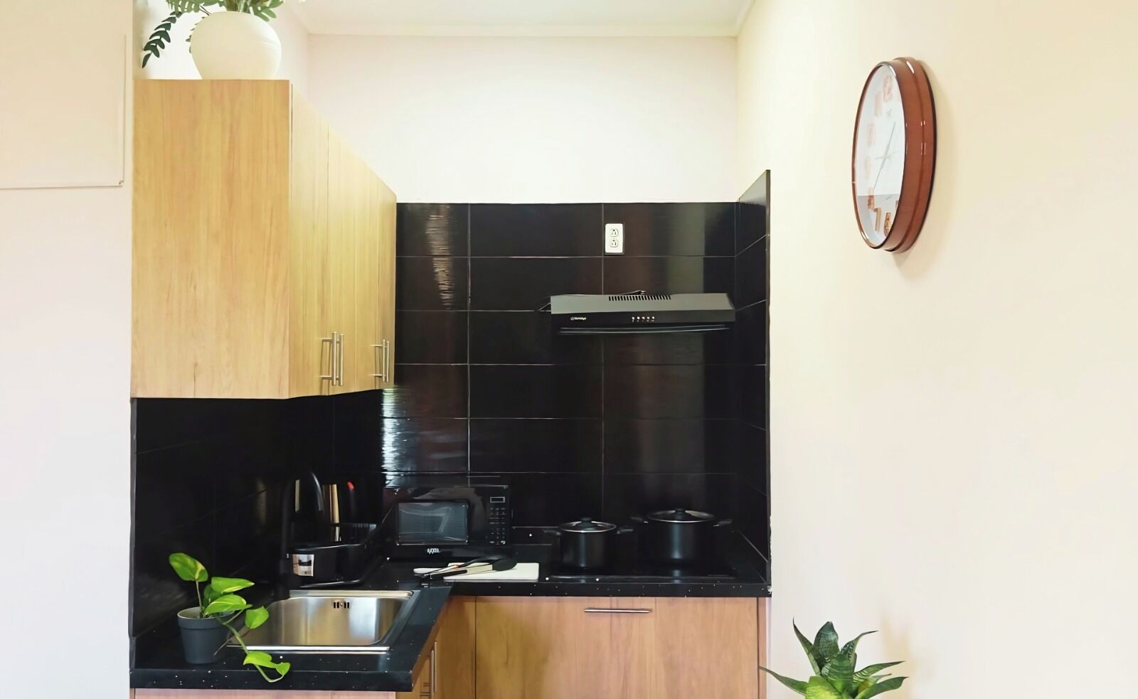 A compact kitchen with black tiles, wooden cabinets, a microwave, a stove, and a clock on the wall.