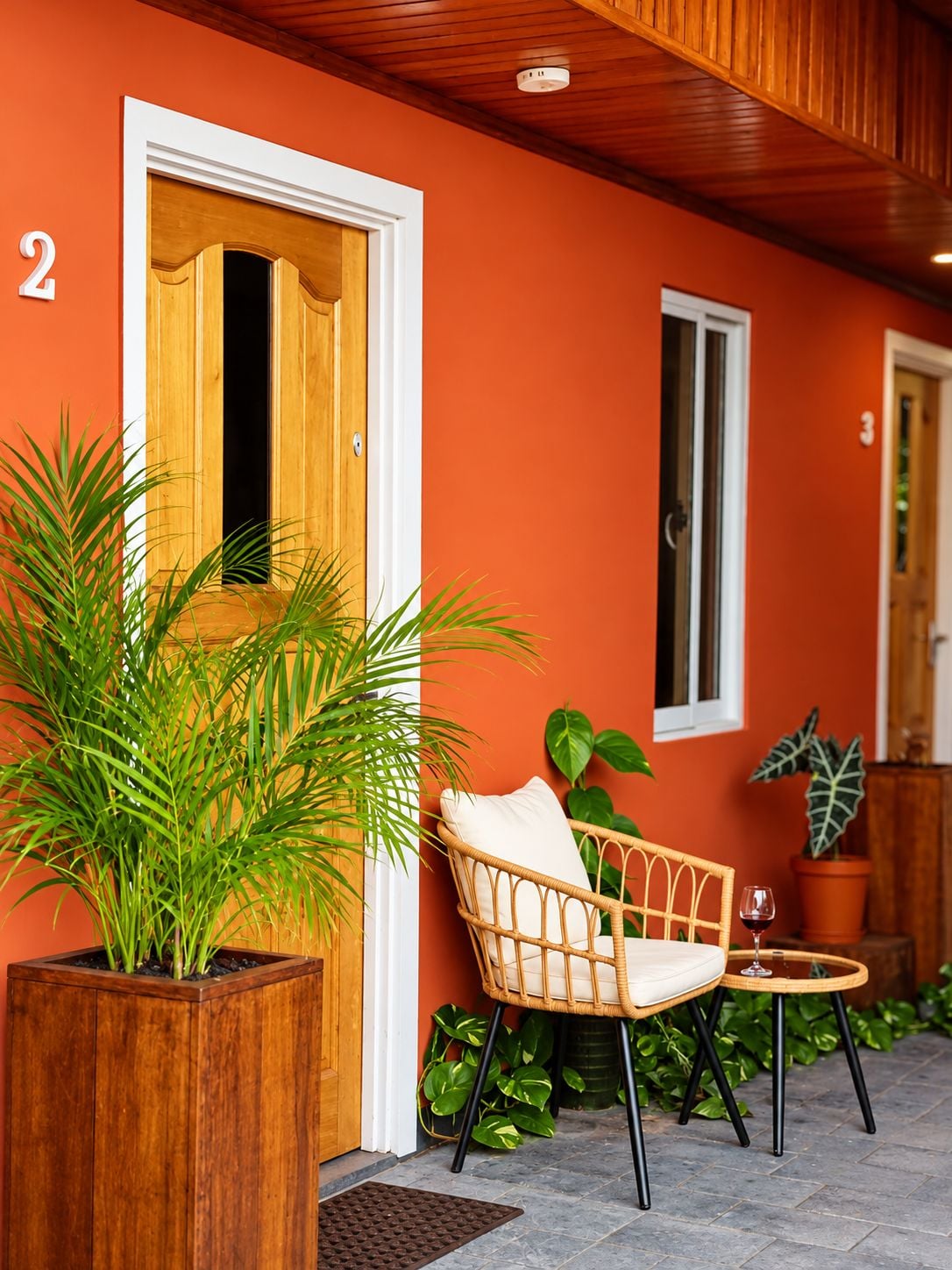 A room with a wooden door, a wicker chair with a cushion, a small table with a glass, and a potted plant.
