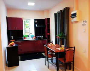 Une cuisine de taille réduite avec des meubles de rangement rouge foncé, un plan de travail noir et un coin repas comprenant une table et des chaises en bois.