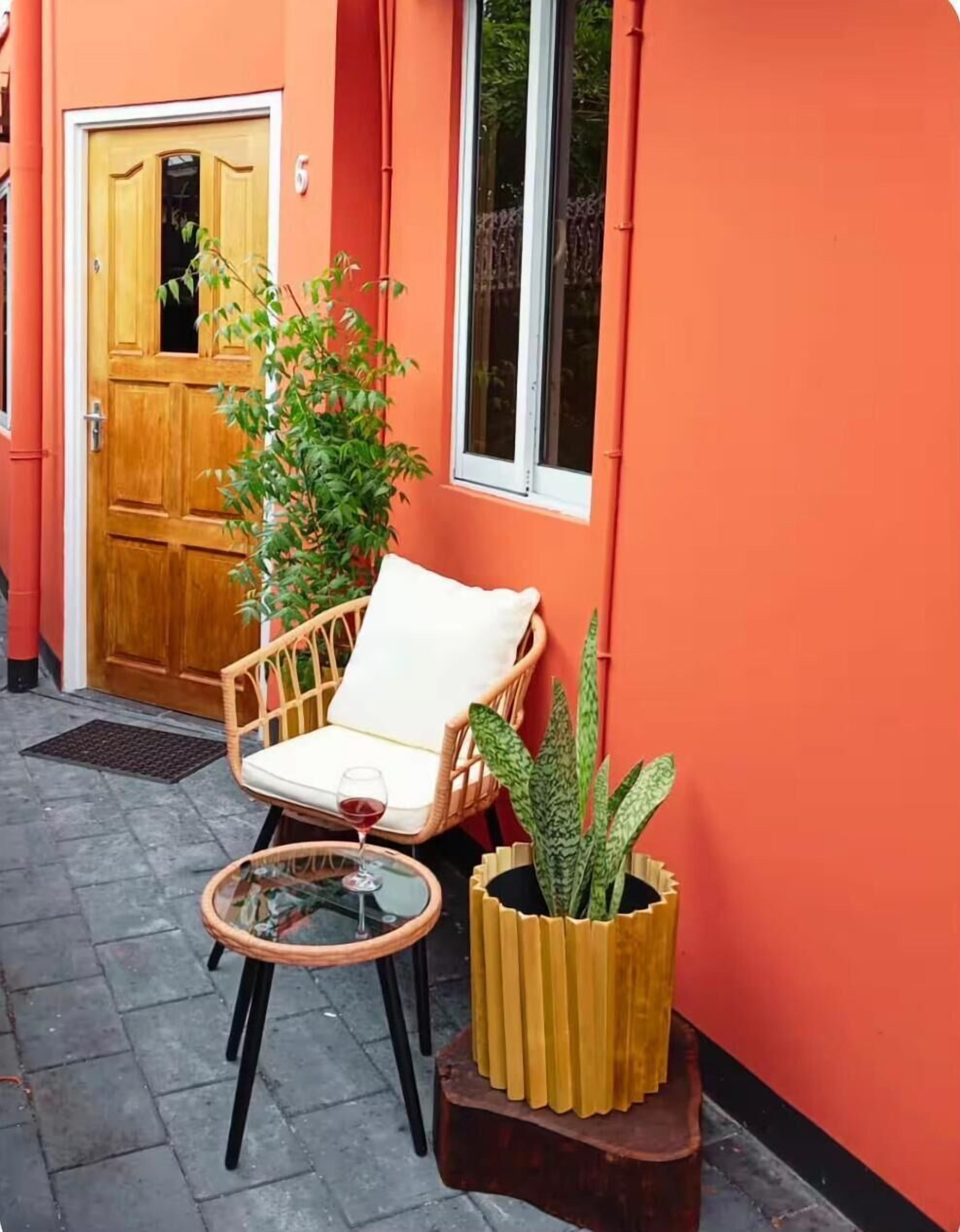 A red building with a wooden door, a wicker chair with a cushion, a small round table with a glass top, and a potted plant.