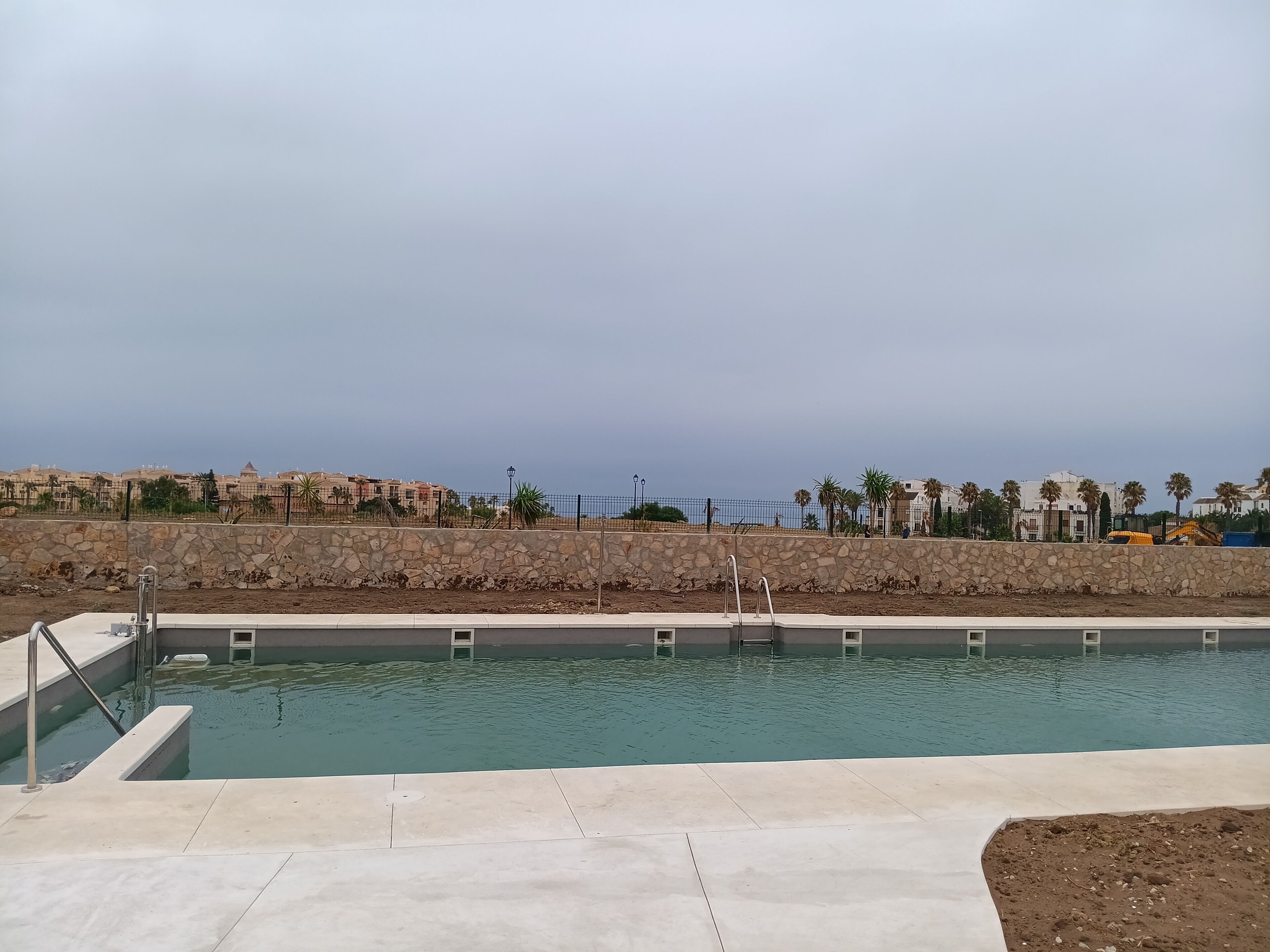 Une piscine bordée d’un mur en pierre, avec en arrière-plan des bâtiments et des palmiers.