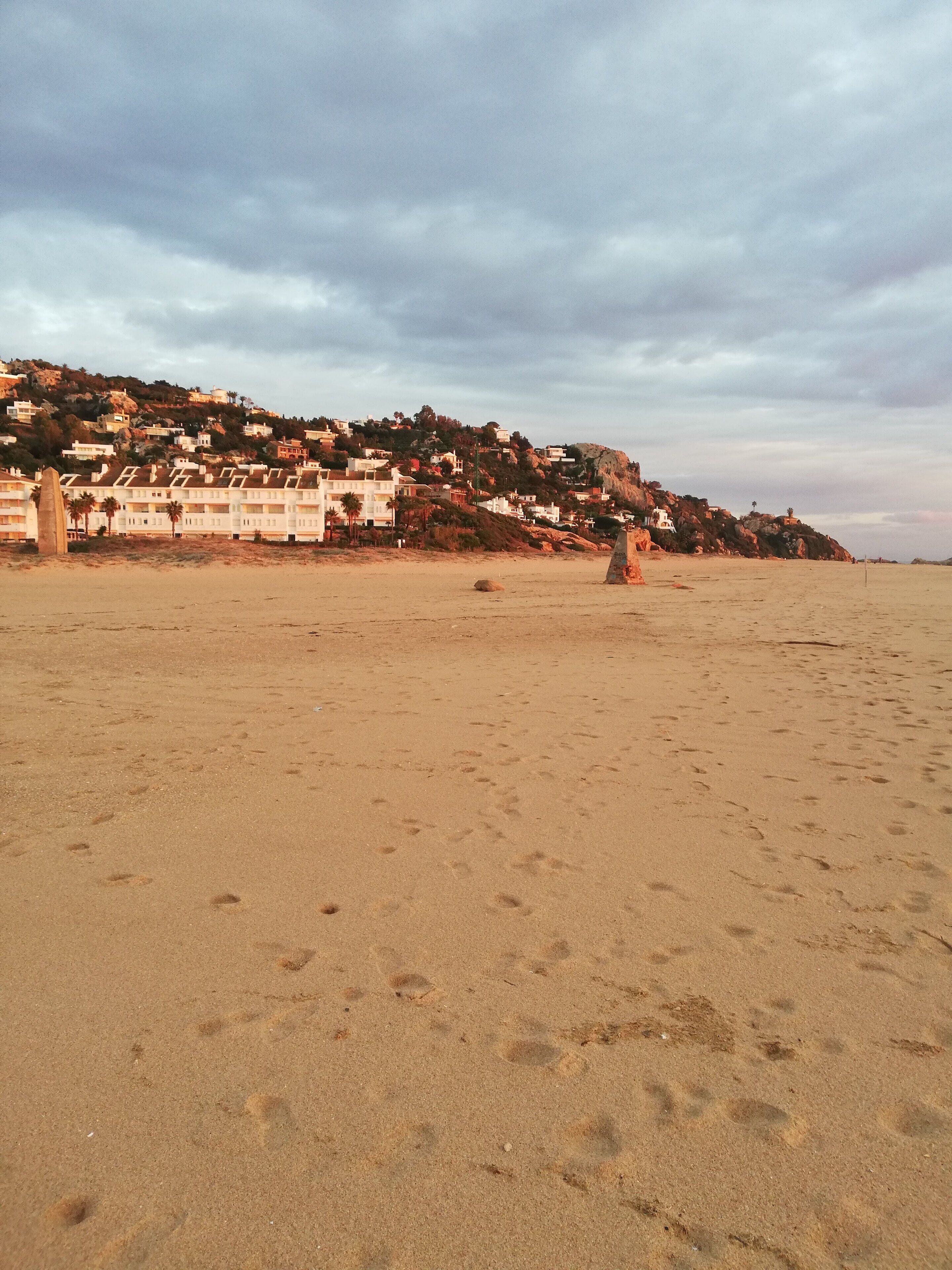 A sandy beach with footprints, a rocky outcrop, and a hillside with buildings.