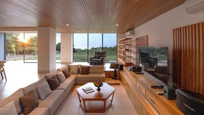 Una sala de estar moderna con techo de madera, ventanas amplias, un sofá, una mesa de centro, un televisor y una estantería.