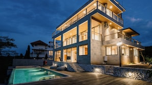 Una casa moderna de varios pisos con piscina, amplias ventanas y un exterior bien iluminado al anochecer.