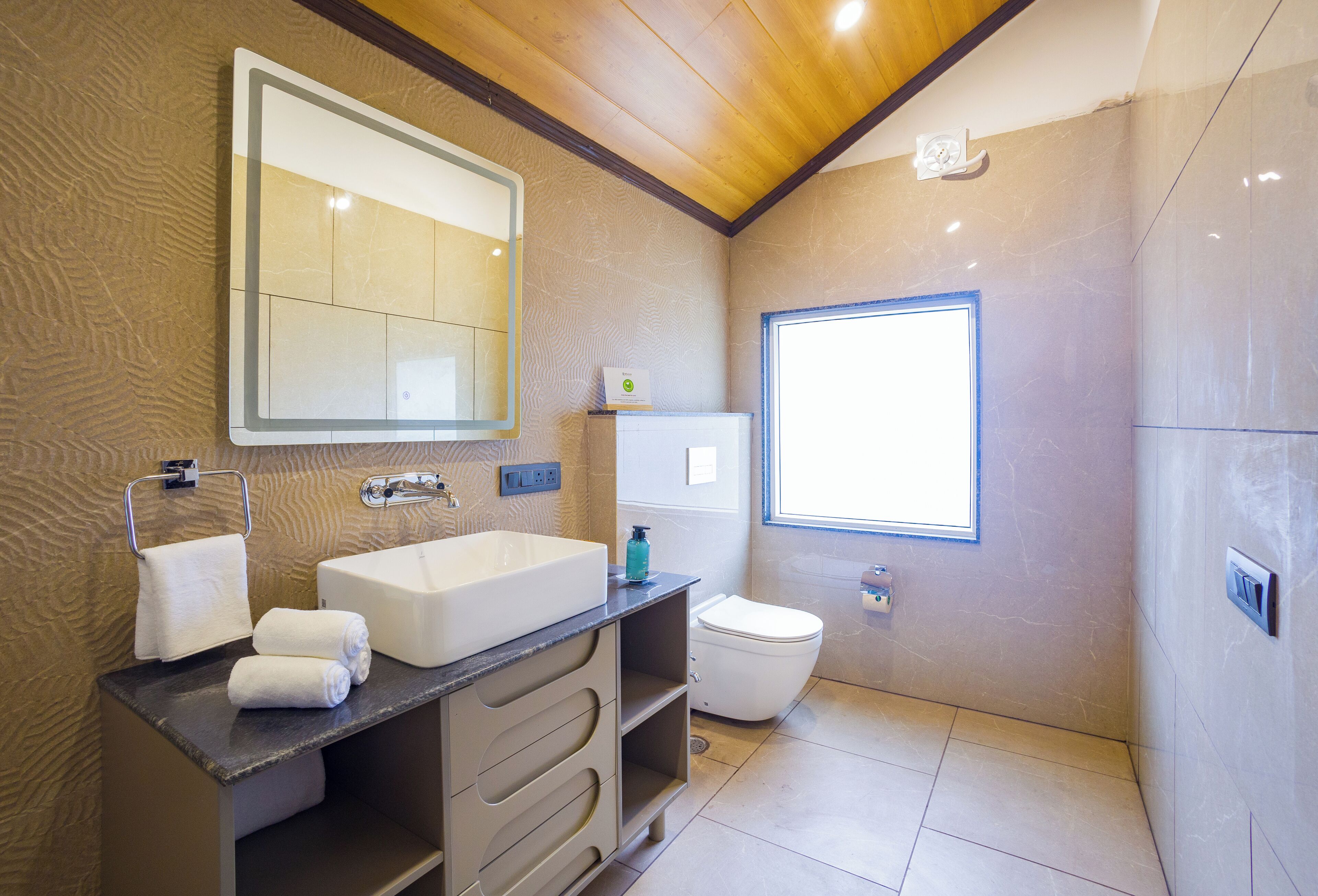 A modern bathroom with a large mirror, a sink, a toilet, and a window providing natural light.