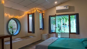 Quarto com cama, ventoinha no teto, janela com cortinas, mesa pequena e ar condicionado fixado na parede.