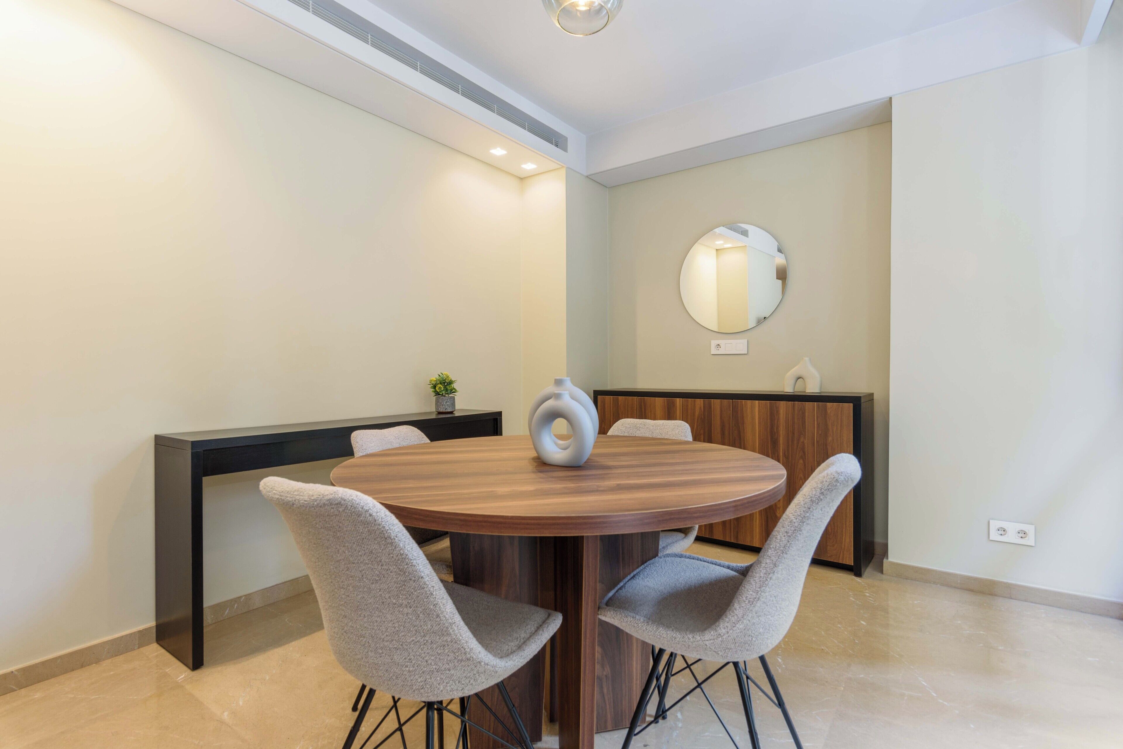 A modern dining area with a round wooden table, two grey upholstered chairs, a black console table, and a round mirror on the wall.
