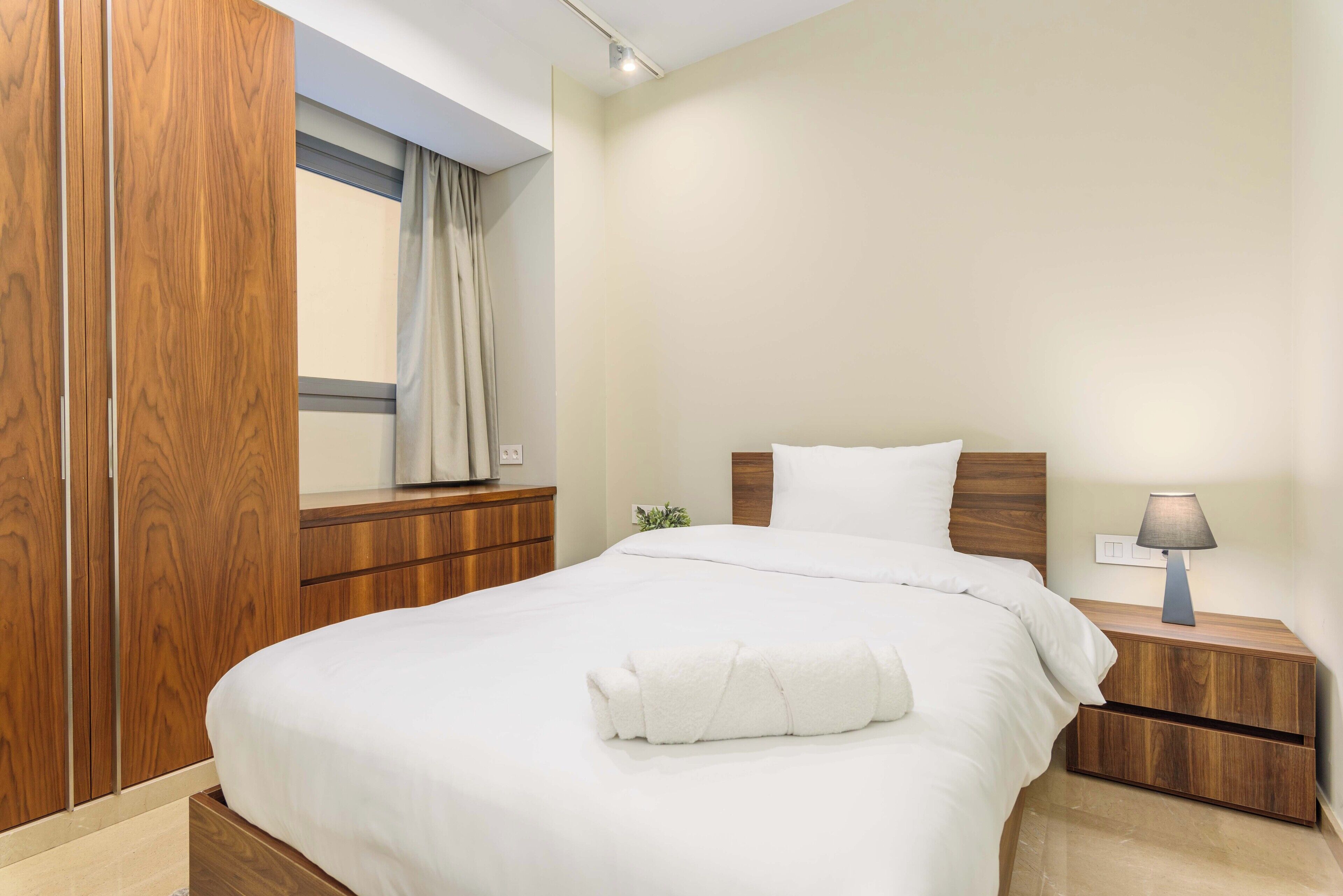 A neatly made bed with a white duvet and a rolled white towel, a wooden nightstand with a lamp, and a wooden wardrobe with a window above it.