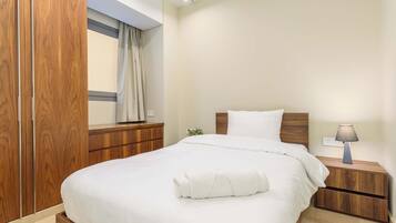 A neatly made bed with a white duvet and a rolled white towel, a wooden nightstand with a lamp, and a wooden wardrobe with a window above it.