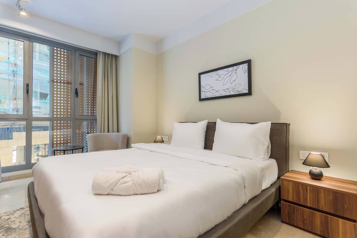 Ein Hotelzimmer mit einem großen Bett, einem Nachttisch mit Lampe, einem Sessel und einem Fenster mit Blick auf Gebäude.