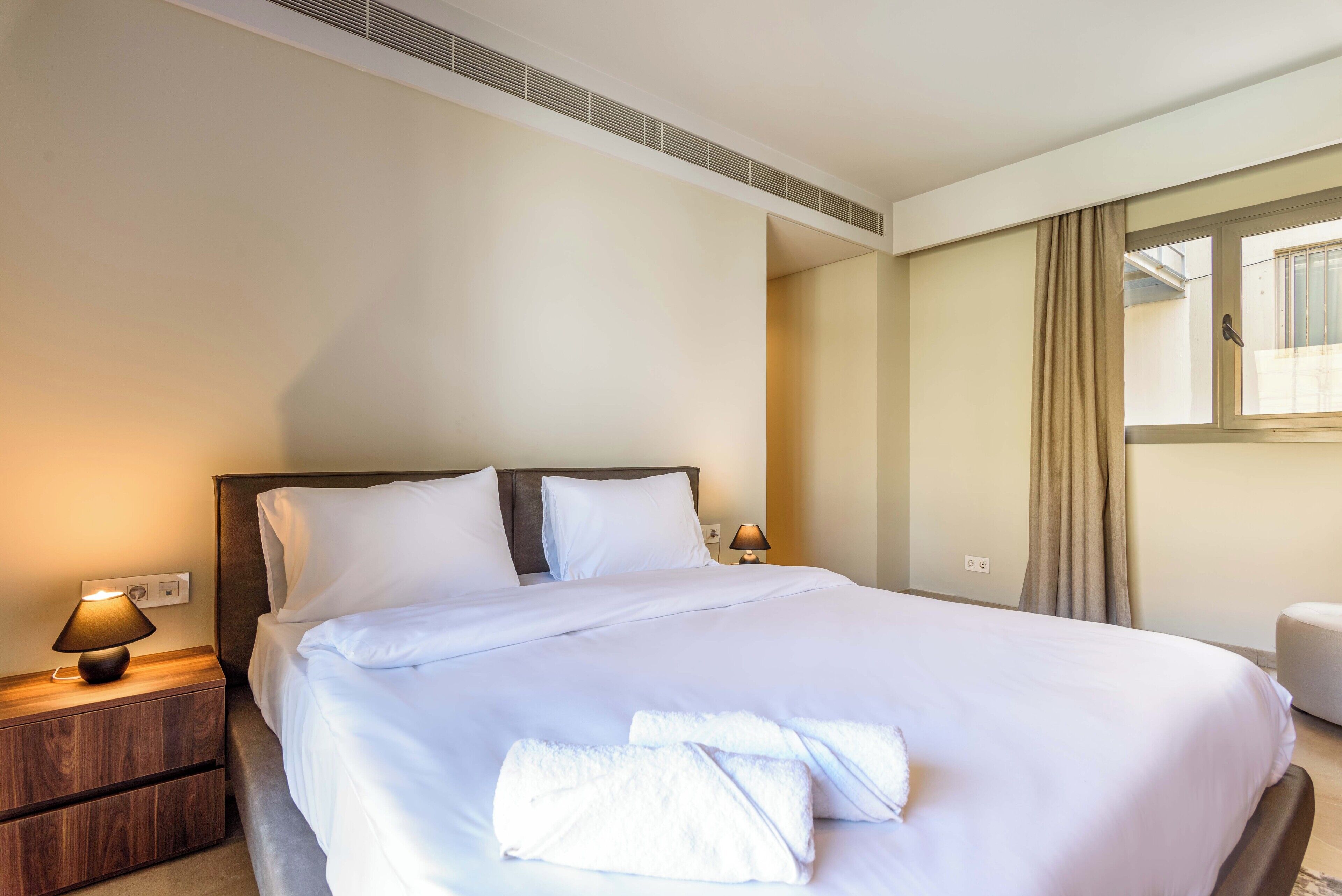 A neatly made bed with white linens and towels, a bedside lamp, and a wooden nightstand.