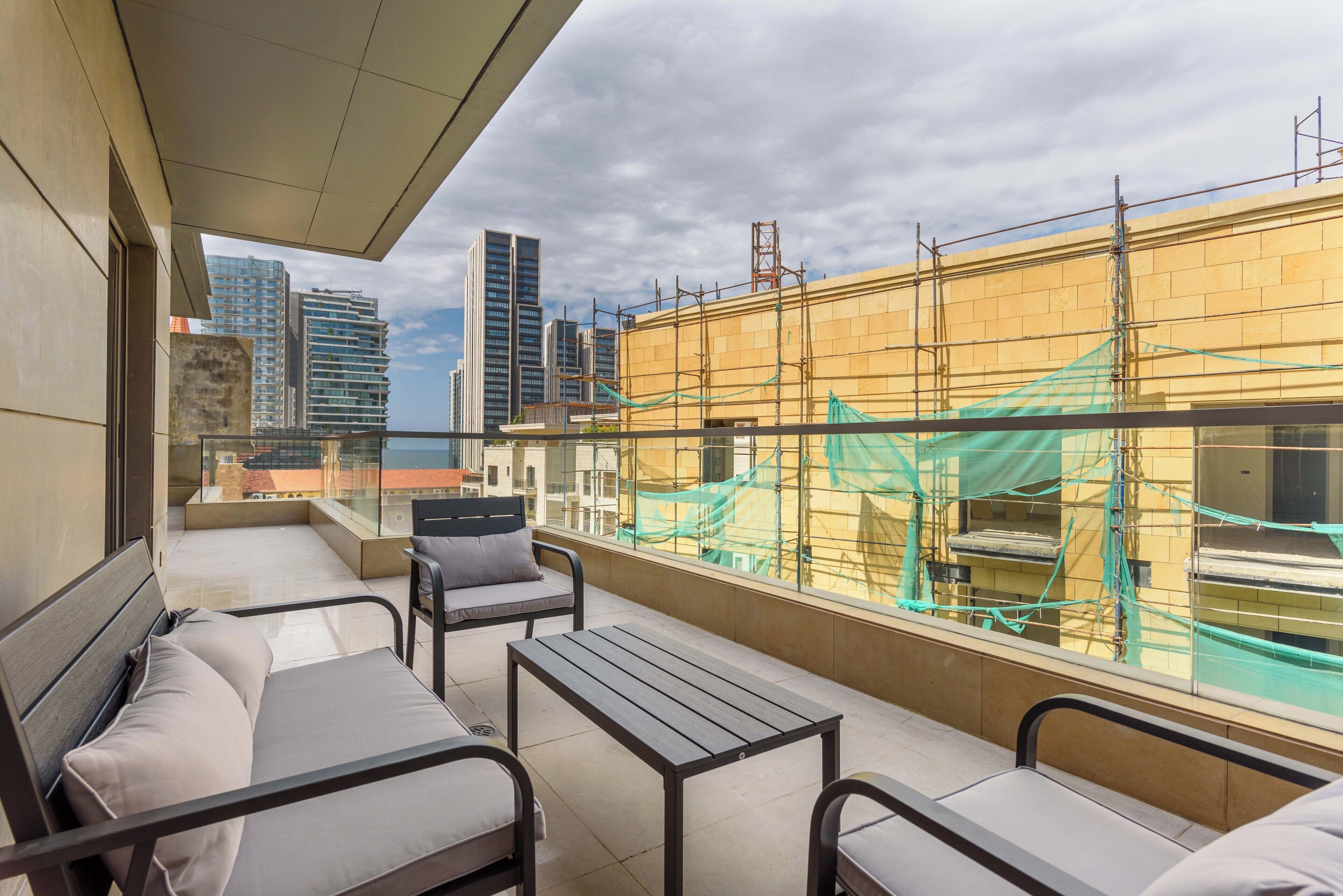 A balcony with a sofa, chairs, and a table, overlooking a building under construction with scaffolding.
