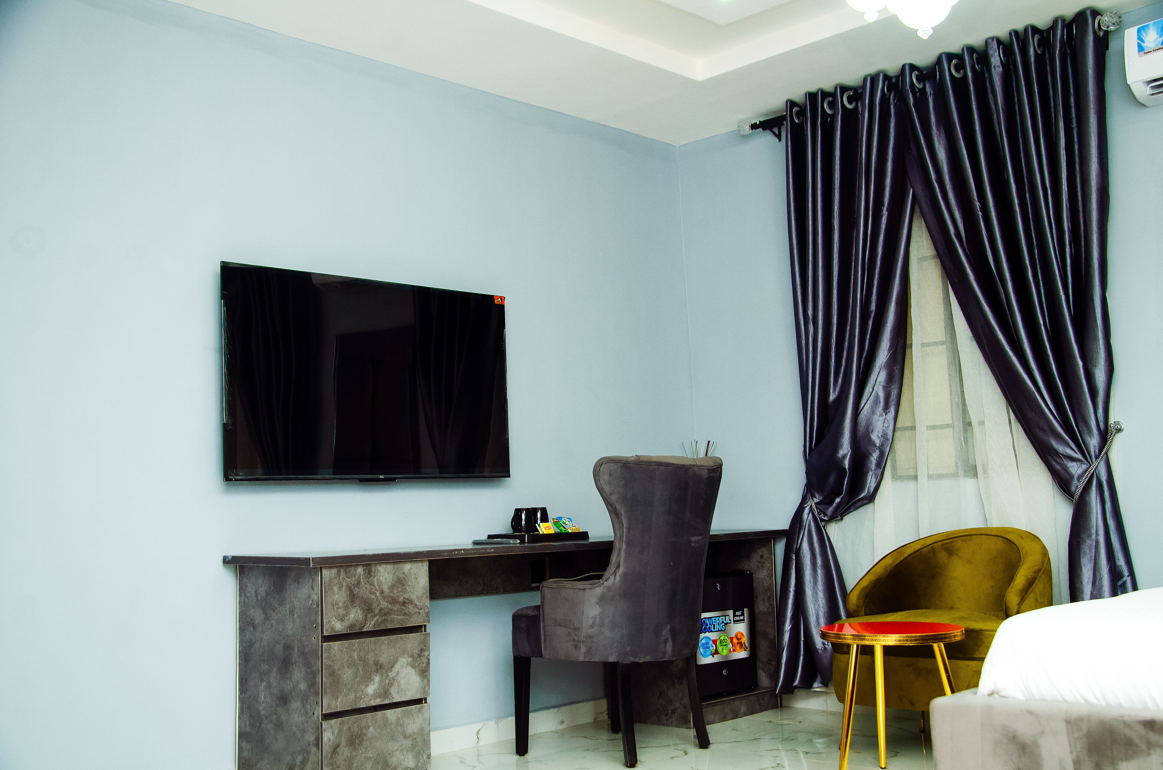 A room with a flat-screen TV mounted on the wall, a desk with drawers, a chair, a small round table with a red top, and a bed with white bedding.
