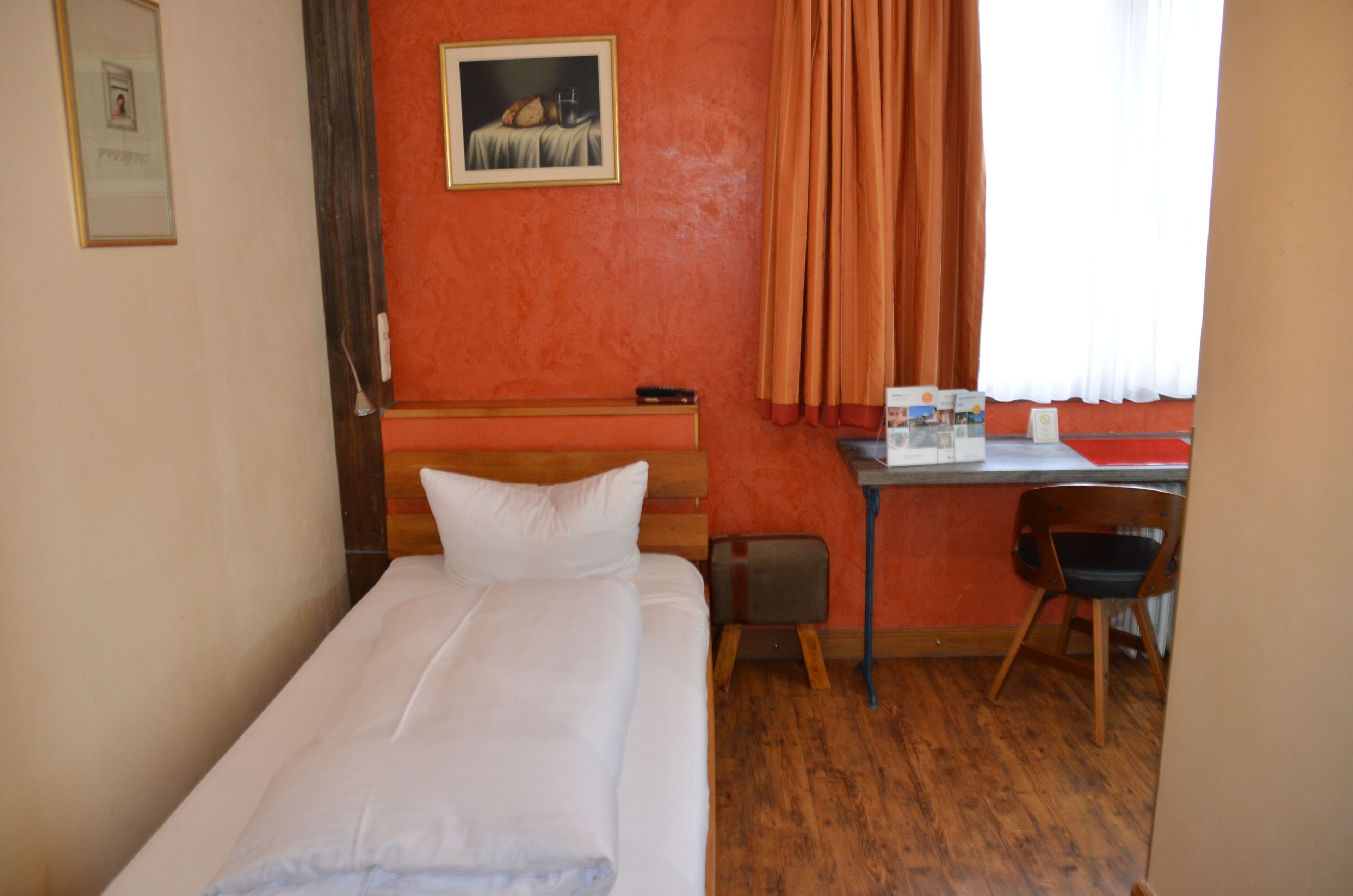 A hotel room with a bed, a desk, a chair, a painting on the wall, and a window with curtains.