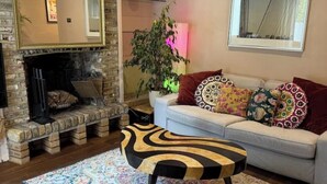 Un salon chaleureux avec une cheminée, un canapé blanc agrémenté de coussins colorés, un tapis à motifs et un pouf original aux couleurs noir et jaune.