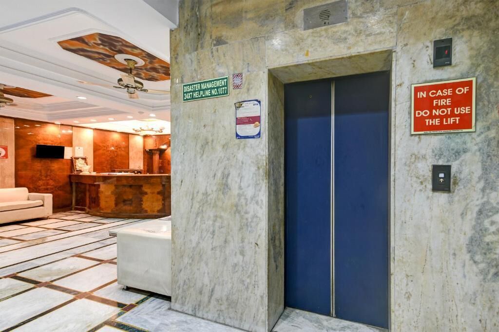 A hotel lobby with a reception desk, a blue elevator, and signs indicating a 24-hour helpline and fire safety instructions.